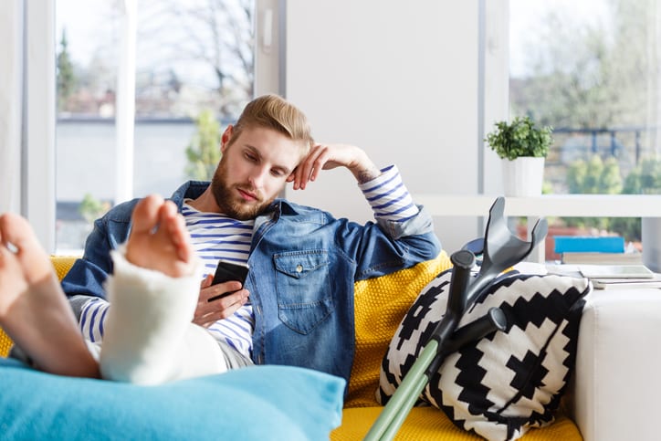 Law Office of Tipton-Downie | Attorney at Law | Vidalia, GA | personal injury Young man with broken leg in plaster cast lying down on sofa at home and using a smart phone